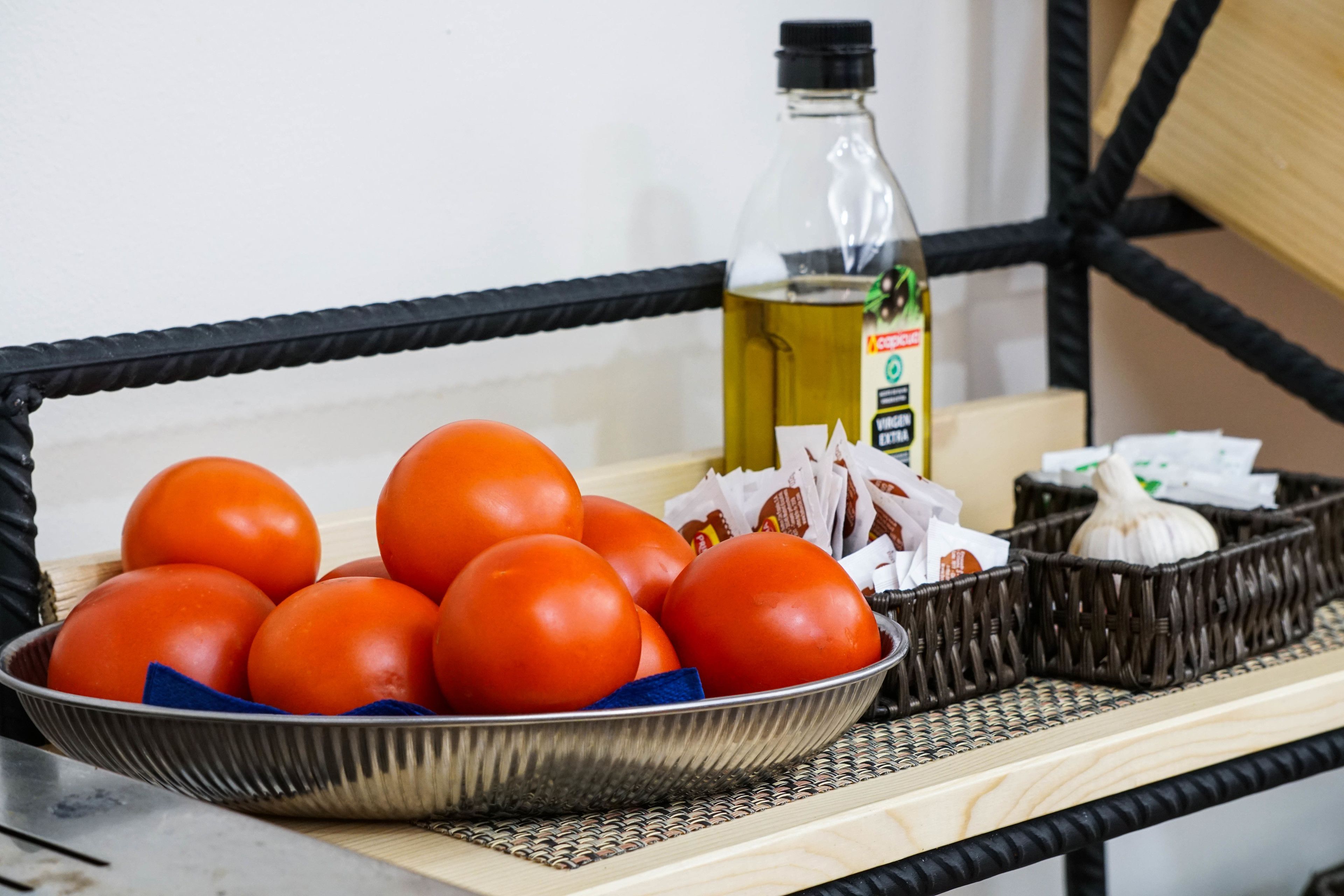 Una cesta de mimbre con tomates, una botella de aceite y dientes de ajo sobre un tapete tejido.