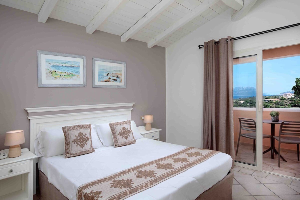 A bedroom with a bed, two bedside tables, a nightstand, a balcony with chairs, and a view of the sea.