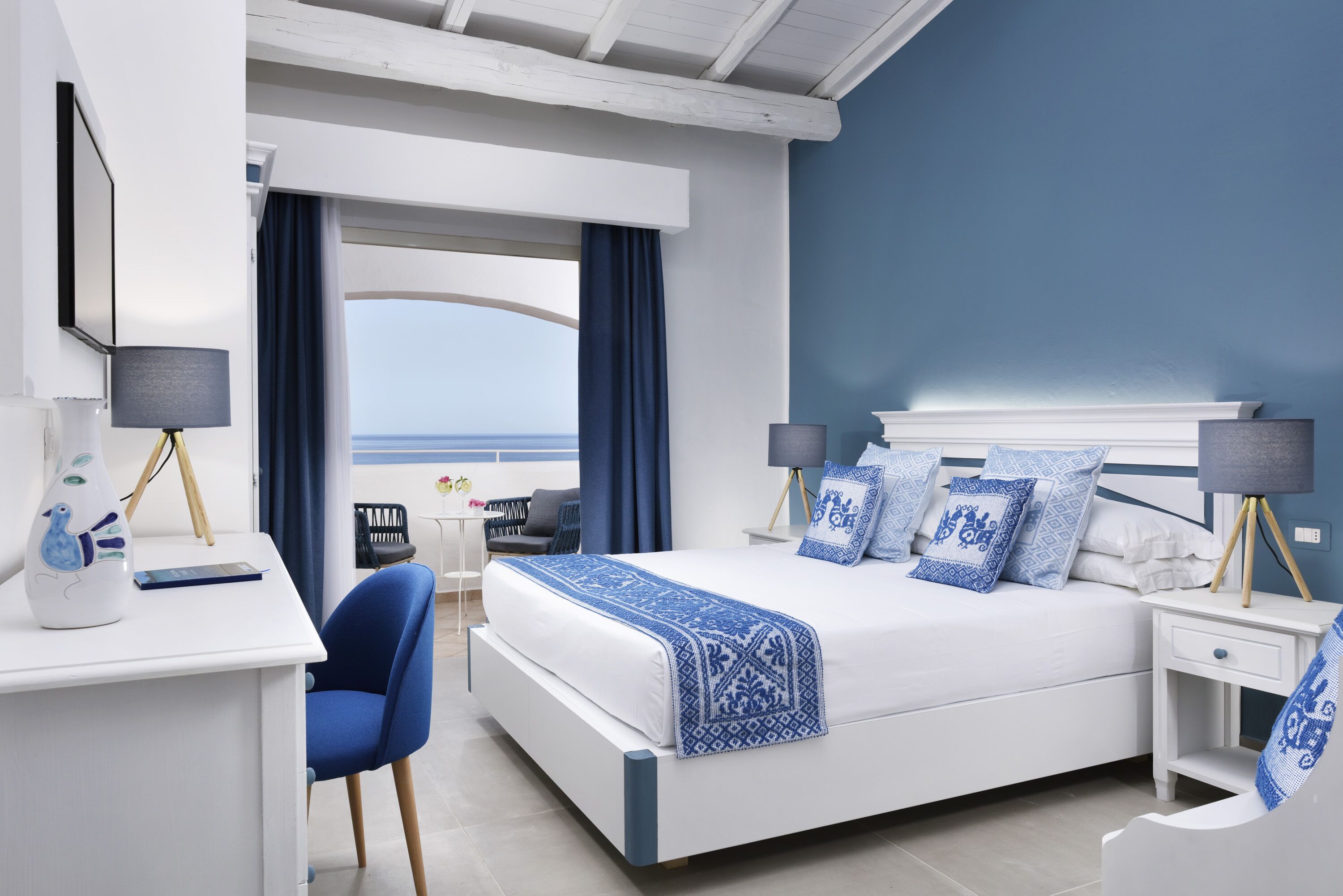 A modern bedroom with a large bed, a desk, and a view of the sea.