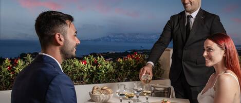 A couple dining at an outdoor table with a view of the sea and mountains.