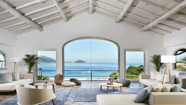 A spacious living room with a large arched window overlooking a beach and ocean. There are comfortable seating areas with sofas and a rocking chair. - VOI Colonna Village (Golfo Aranci)
