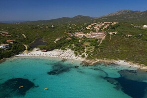 An aerial view of a coastal resort with a beach, clear turquoise waters, and a mountainous backdrop. - VOI Colonna Village (Golfo Aranci)