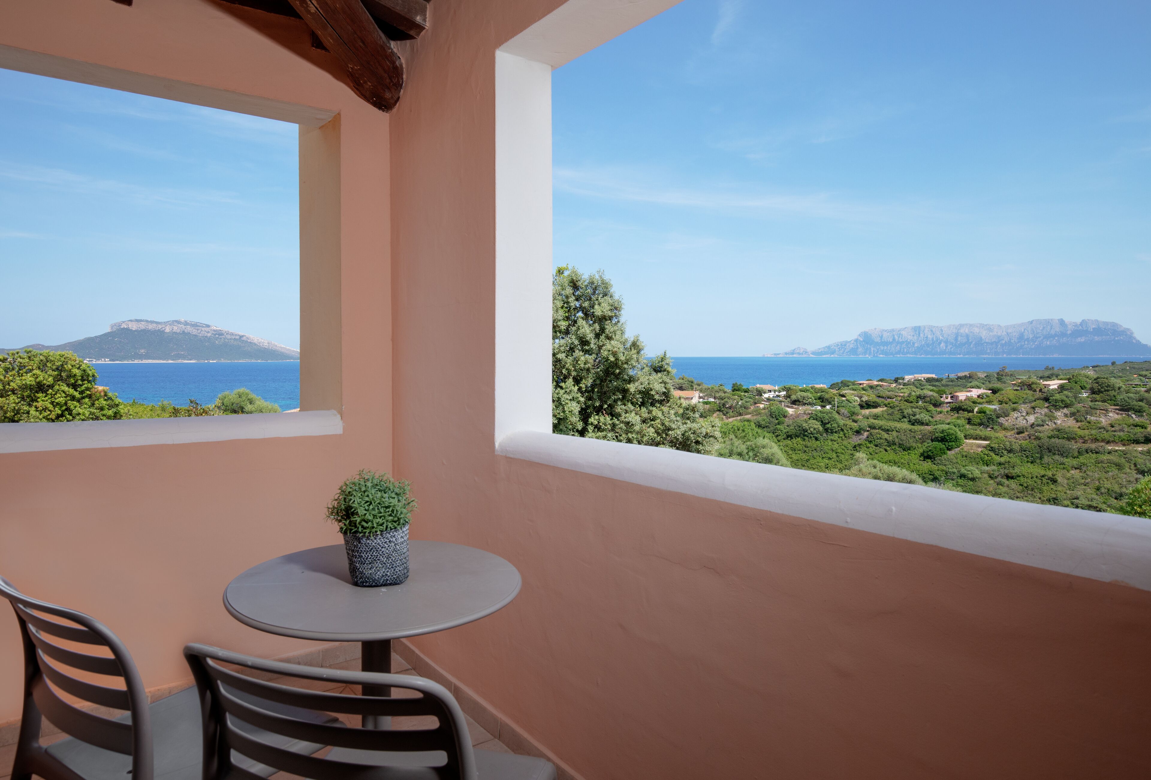 A balcony with a table and chairs, overlooking a coastal landscape with trees and a distant mountain.