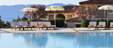 A pool area with lounge chairs and umbrellas, a building with a tiled roof, and a view of the sea.