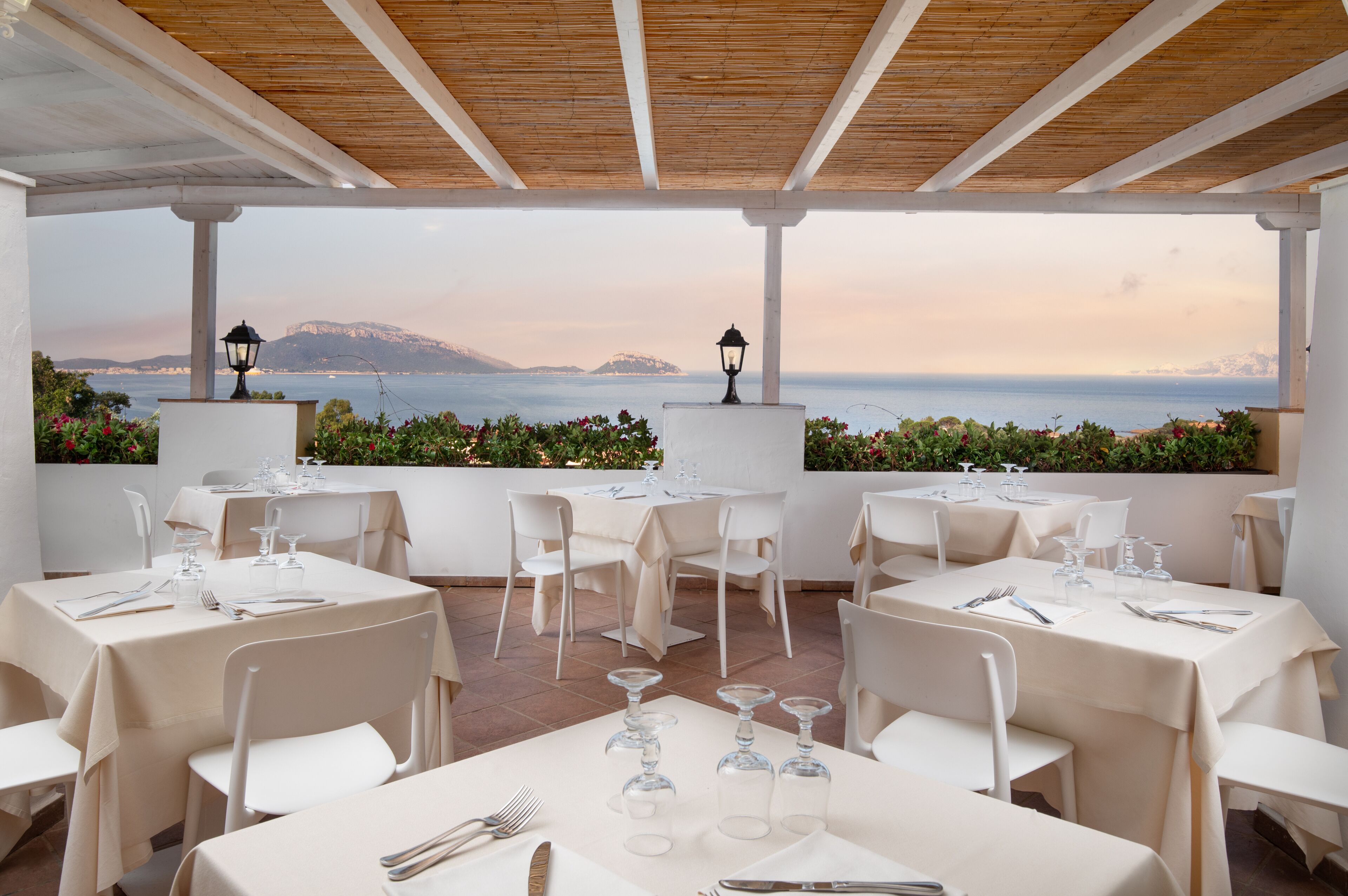 Un restaurant aux tables et chaises blanches, situé dans un cadre offrant une vue magnifique sur la mer et les montagnes.
