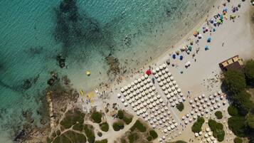 Vista aérea de una playa con hileras de sombrillas y sillas de playa, costa rocosa y agua turquesa cristalina.
