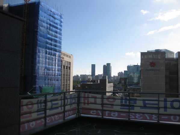 A cityscape with a building under construction covered in blue netting, a clear sky, and a few other buildings in the background. - Laketel (Seoul)