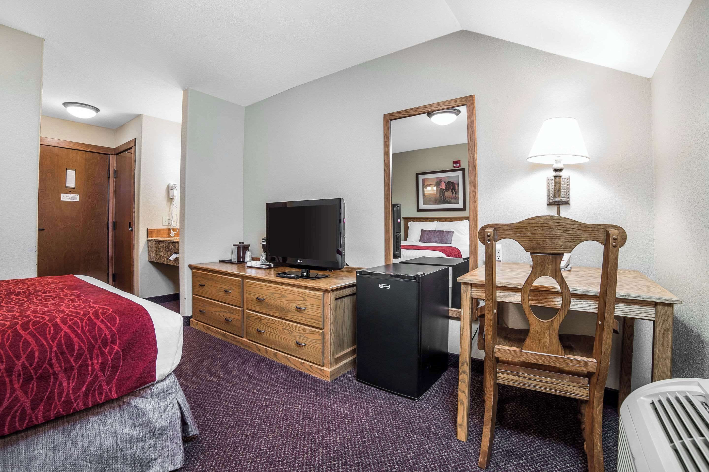 A hotel room with a bed, a desk, a television, a refrigerator, a wooden chair, a mirror, and a lamp.