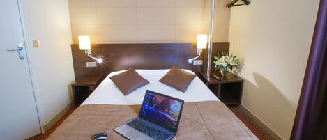 Una habitación de hotel con cama, una computadora portátil, una mesita de noche con lámpara y una mesita auxiliar con planta.