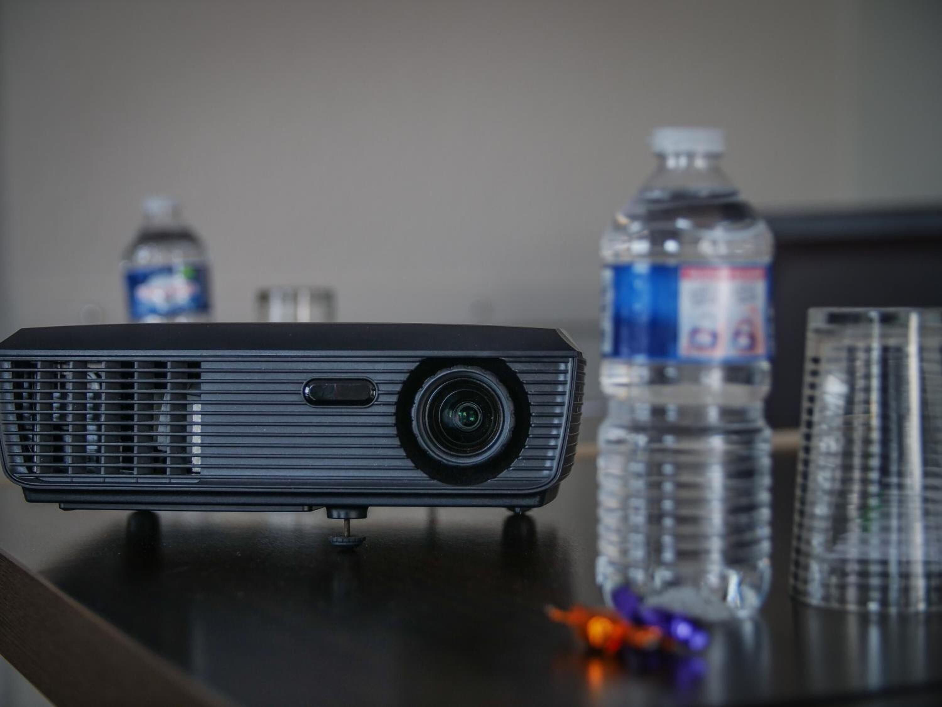A projector on a table with a water bottle and glasses in the background.
