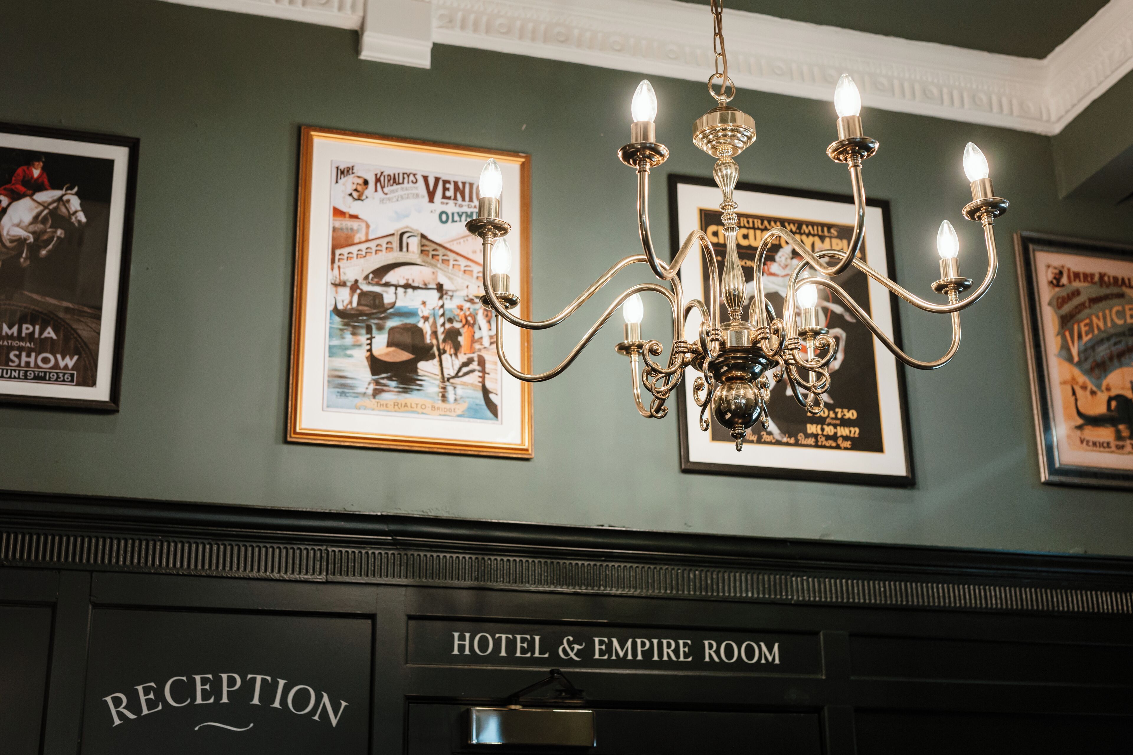 Un área de recepción con una araña, carteles enmarcados y un letrero que indica Hotel & Empire Room.