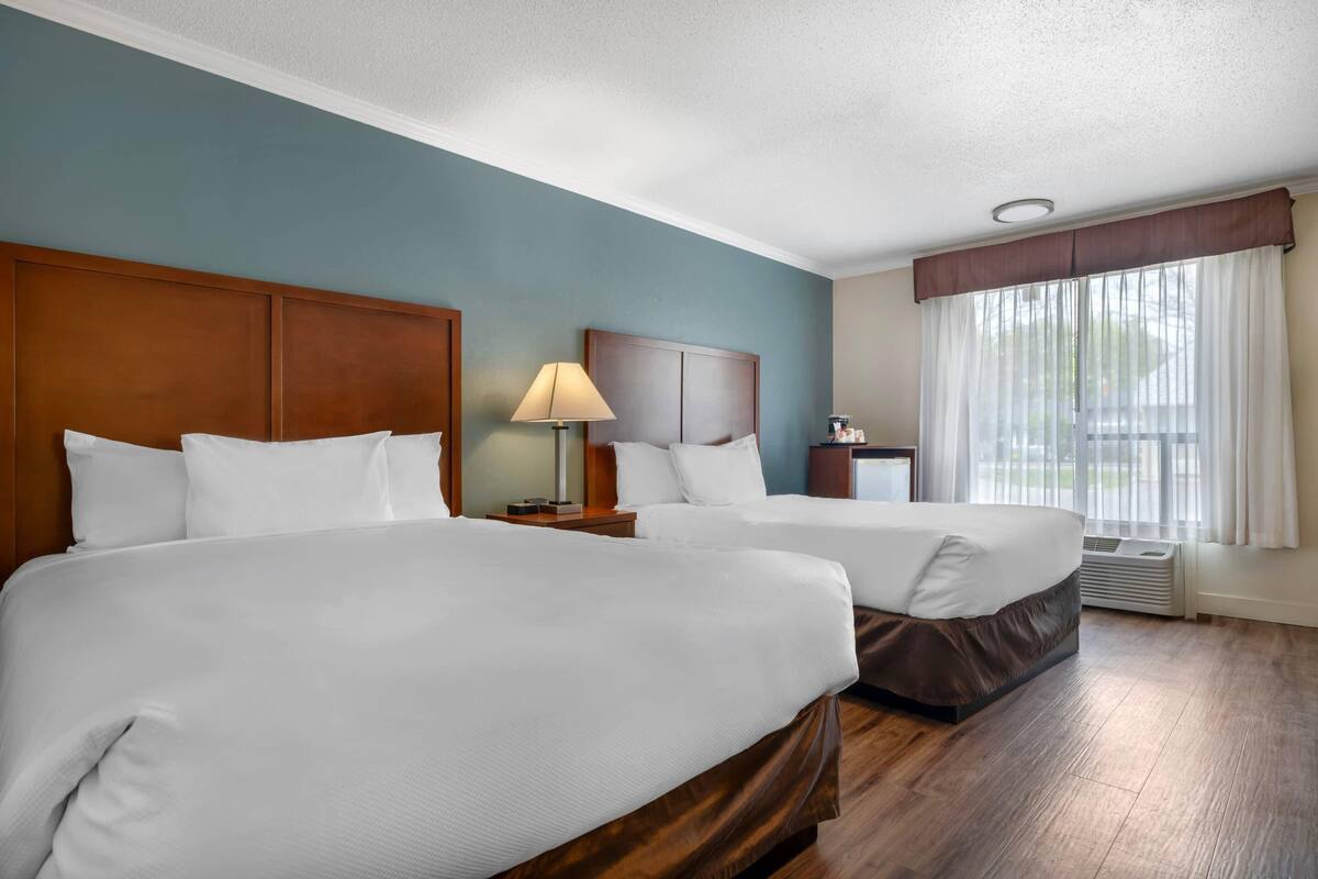 A hotel room with two beds, a nightstand with a lamp, a small table, and a window with curtains.