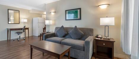 Una sala moderna con un sofá, una mesa de centro, un escritorio con una silla, un refrigerador y un cartel de bienvenida en la pared.