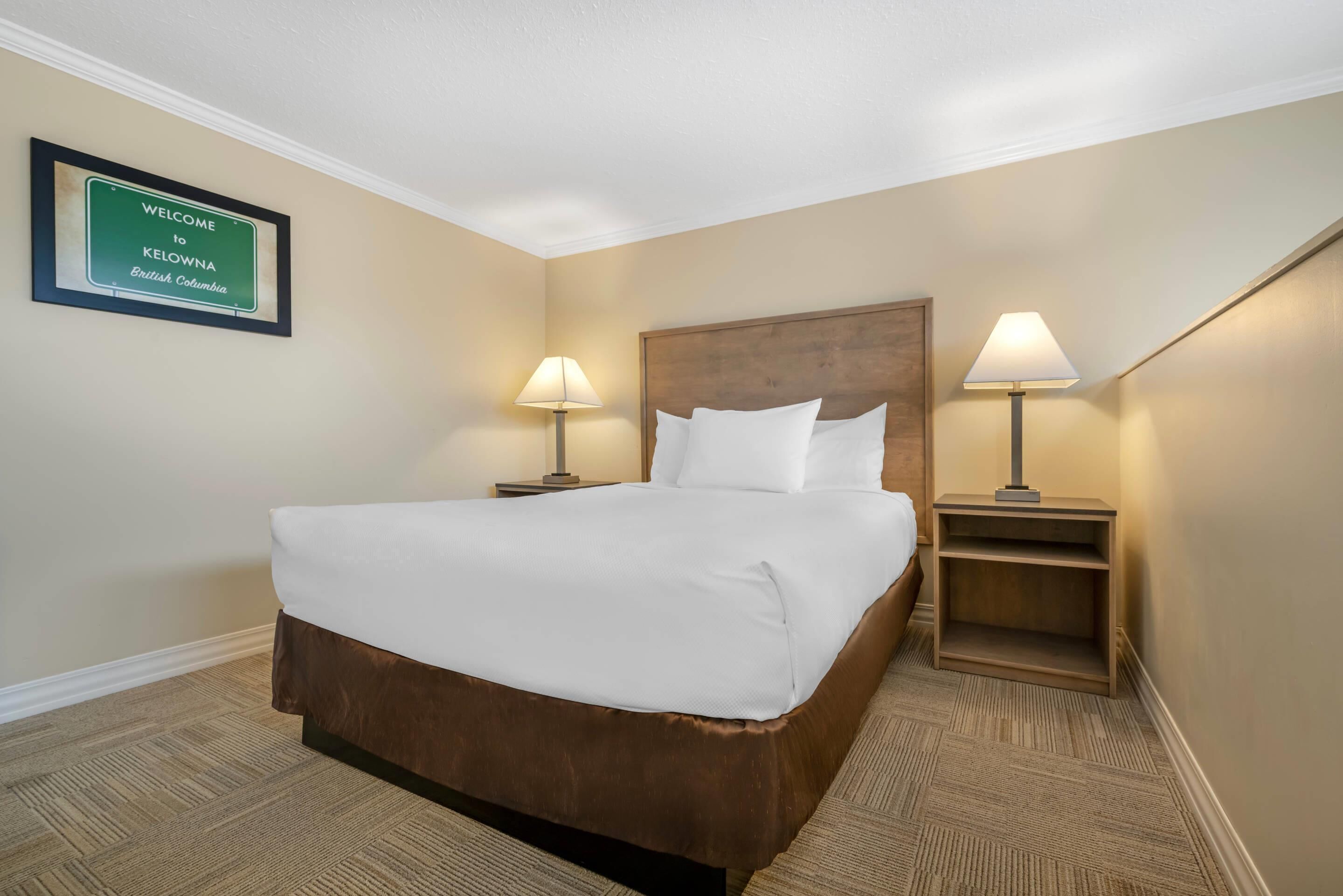 Habitación de hotel con una cama grande, dos mesitas de noche con lámparas, un cartel de bienvenida enmarcado y suelo alfombrado.