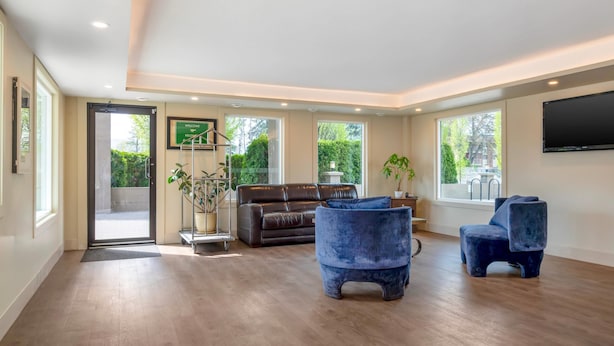 A spacious living area with a brown leather sofa, two blue armchairs, a flat-screen TV, and large windows offering a view of greenery.