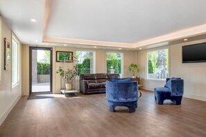 Un amplio salón con un sofá de cuero marrón, dos sillones azules, un televisor de pantalla plana y grandes ventanas con vistas a zonas verdes.