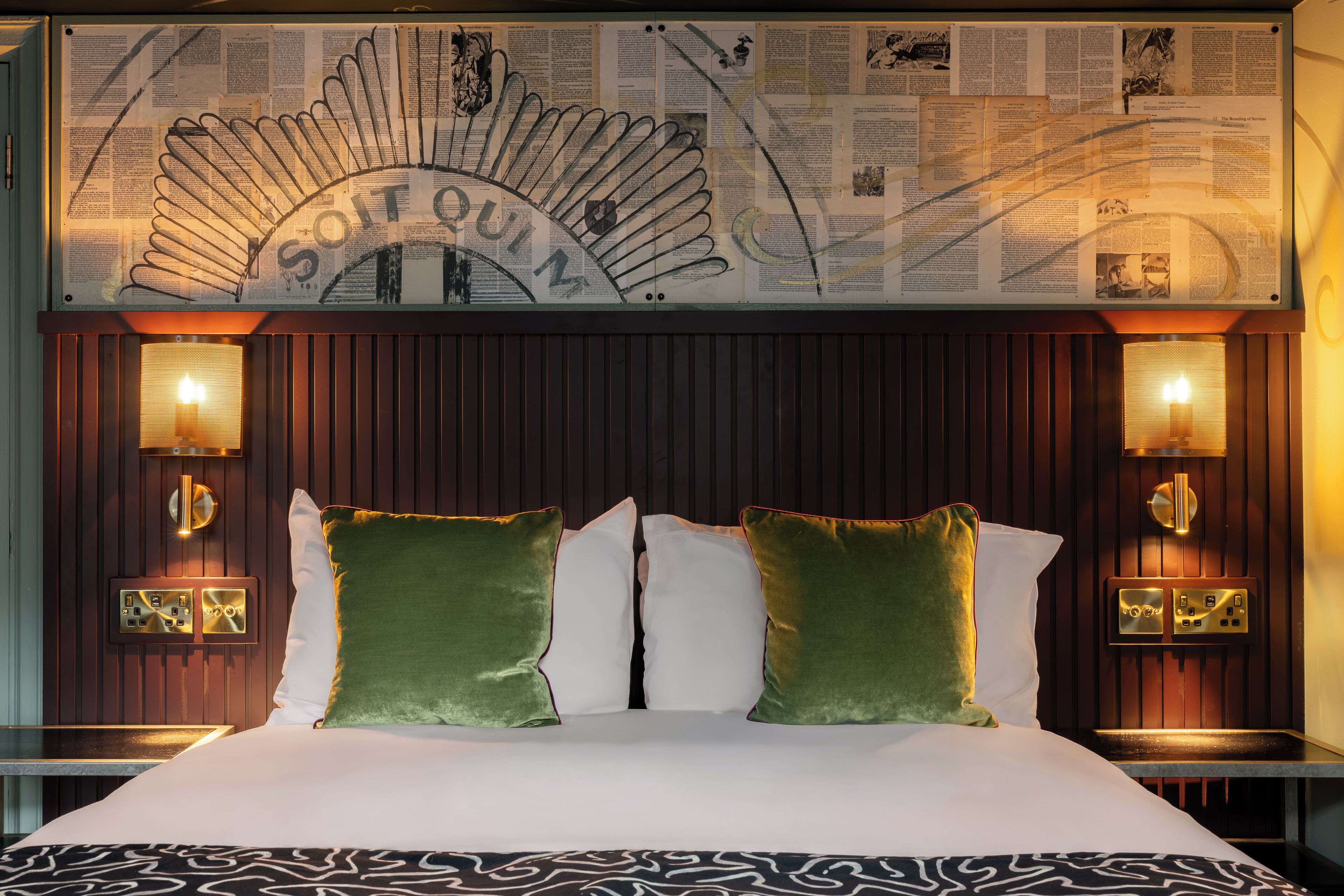 Habitación de hotel con una cama grande, dos almohadas verdes, cabecera con un atril para periódicos y luces de lectura fijadas a la pared.