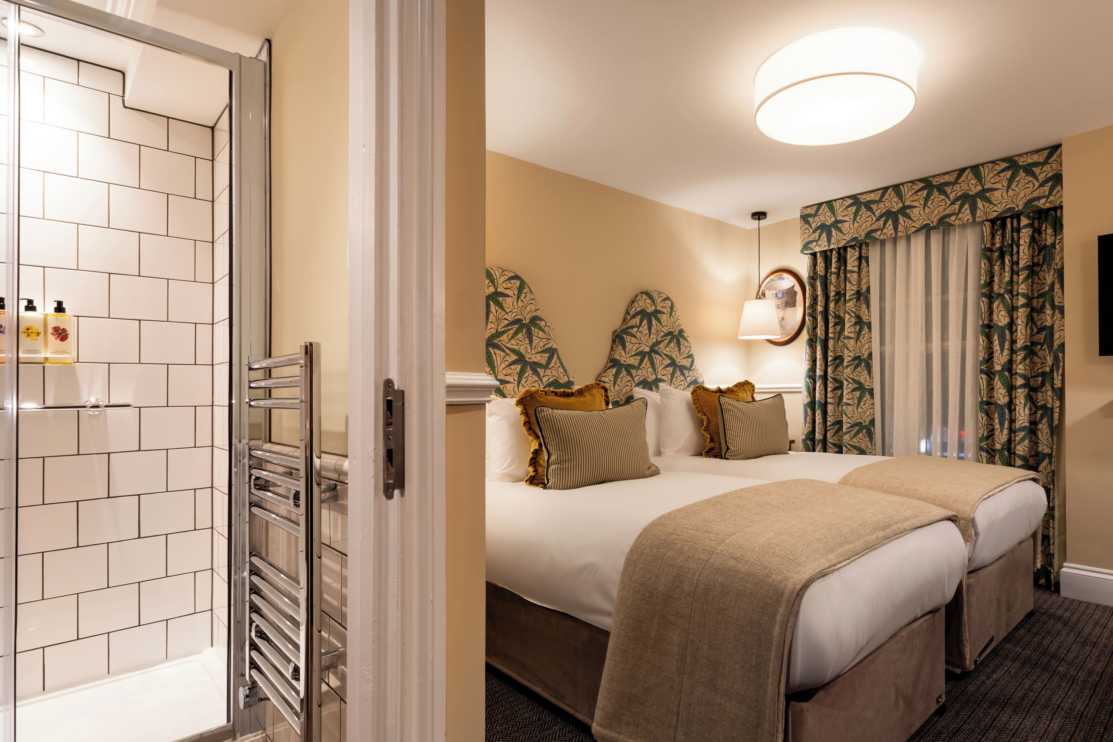 A hotel room with a shower area, a bed with two pillows, a bedside table, and a large window with curtains.