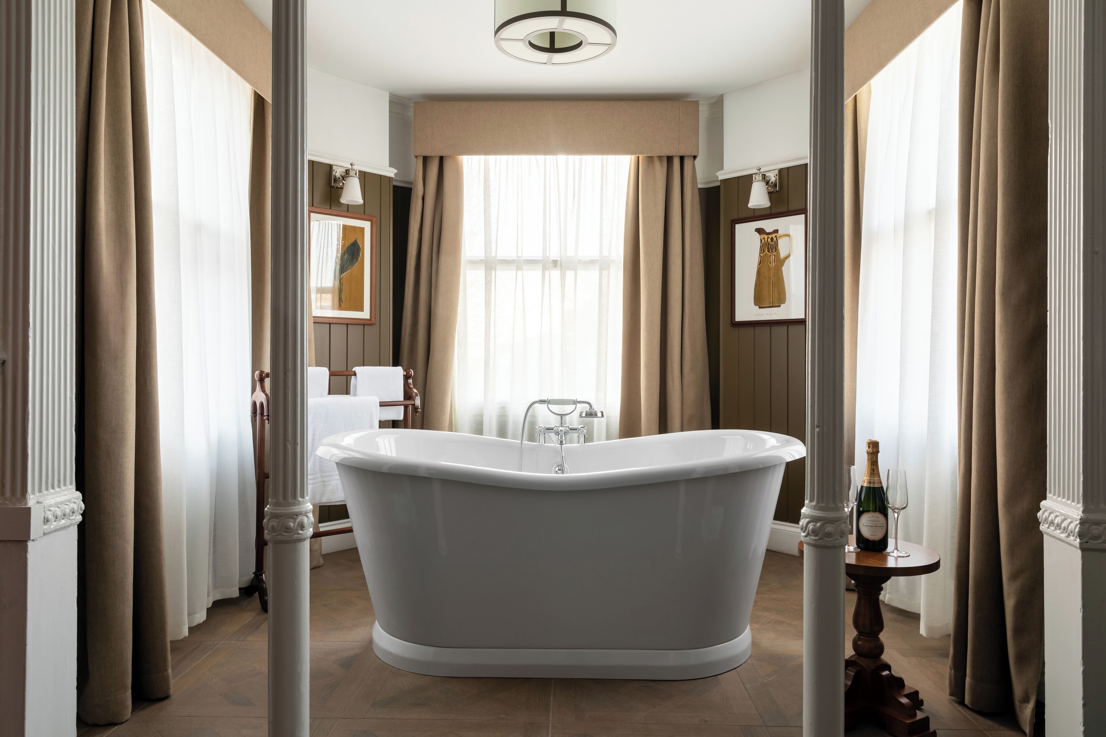 A classic bathtub in a well-lit bathroom with a large window, curtains, and a bottle of champagne on a small table.