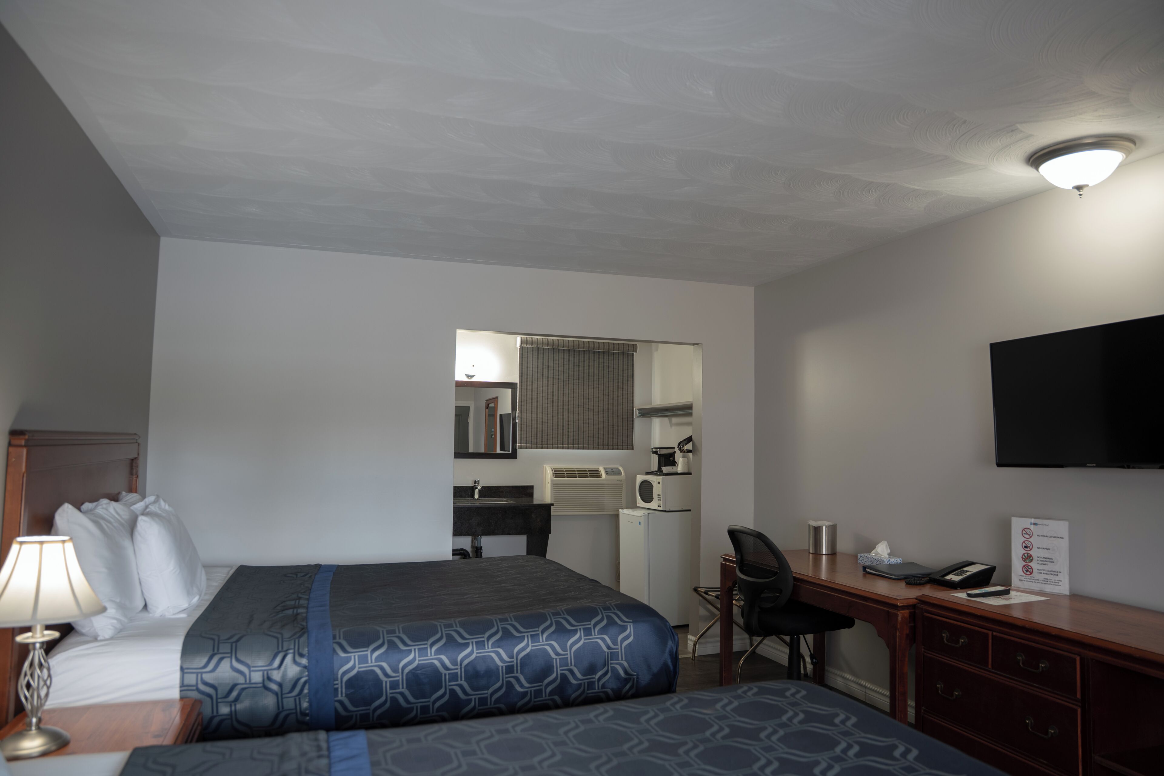 A hotel room with two beds, a desk, a chair, a TV, a small kitchenette, and a mirror.