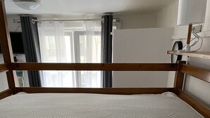 Huoneessa on sänky, puinen pääty, ikkuna, jossa on verhot, ja seinälle kiinnitetty kello.