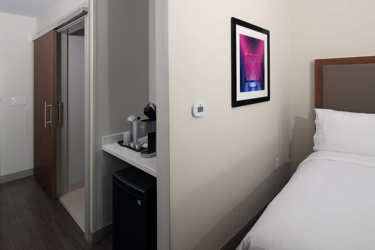 A hotel room with a bed, a coffee machine, a small refrigerator, a wardrobe, and a framed picture on the wall.