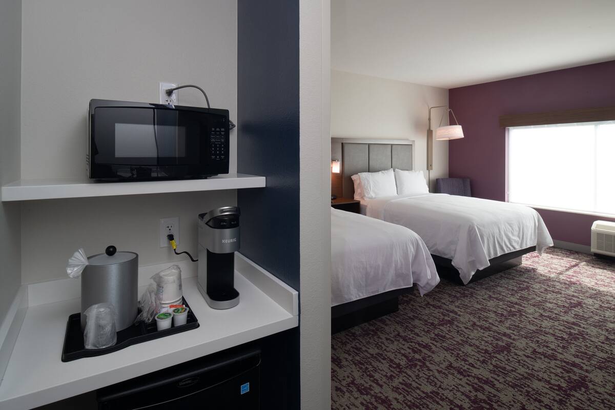 A hotel room with a microwave on a shelf, a coffee maker, and a trash bin. The room has two beds, a nightstand, and a window with a view.