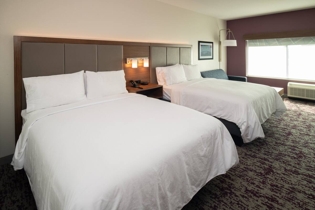 A hotel room with two beds, a nightstand, a lamp, and a window with a view.