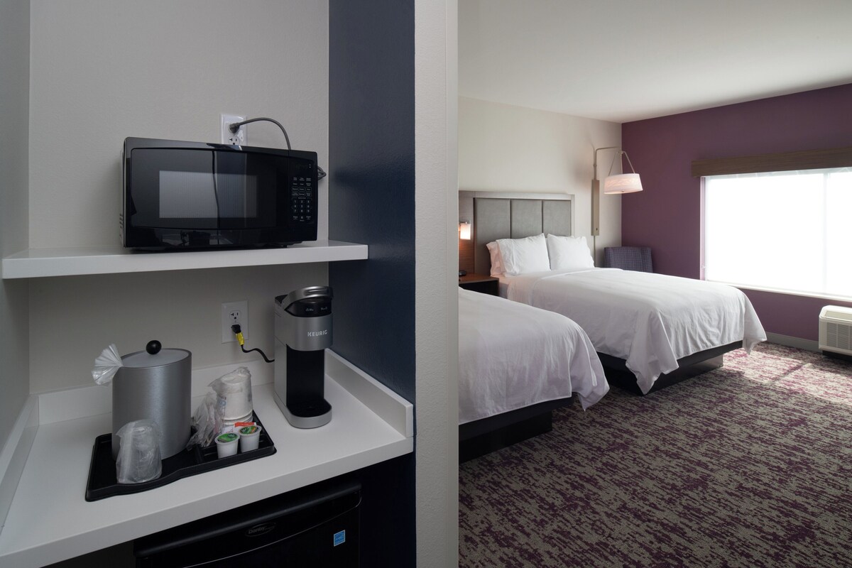 Hotelkamer met een magnetron op een plank, een koffiezetapparaat en een prullenbak. De kamer heeft twee bedden, een nachtkastje en een raam met uitzicht.