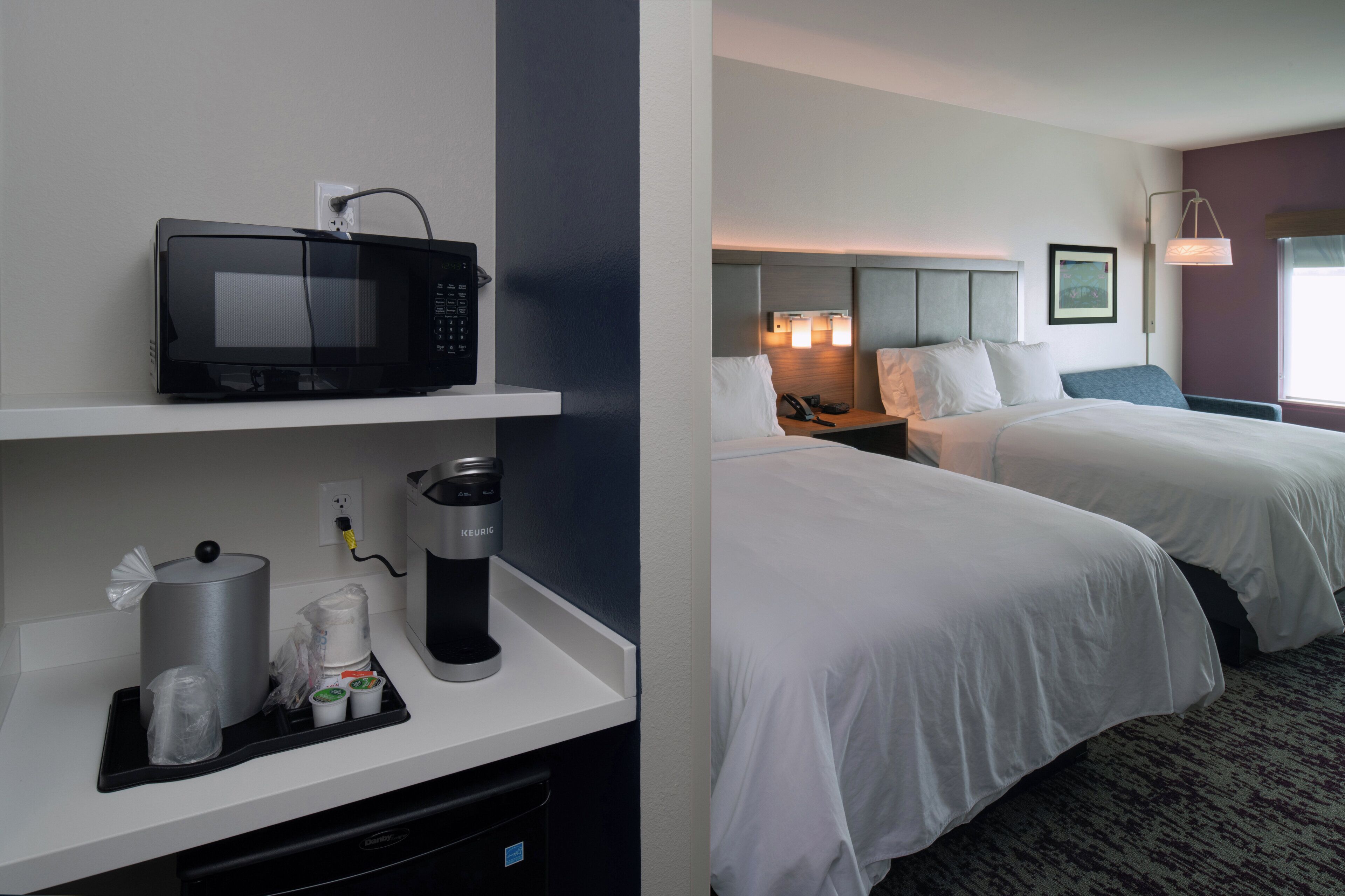 A hotel room with a microwave on a shelf, a coffee maker, and a trash bin. There are two beds with white linens and a nightstand with a lamp.