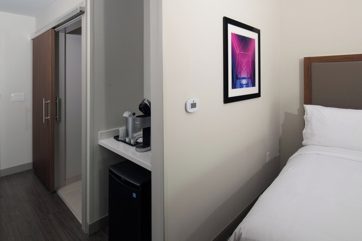 Een hotelkamer met een bed, een koffieapparaat, een kleine koelkast, een kledingkast en een ingelijste foto aan de muur.