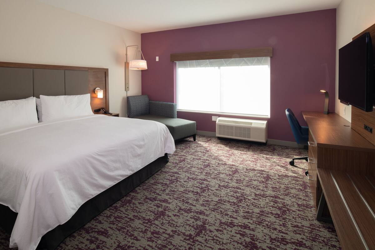 A hotel room with a large bed, a desk, a chair, a TV, and a window with blinds.