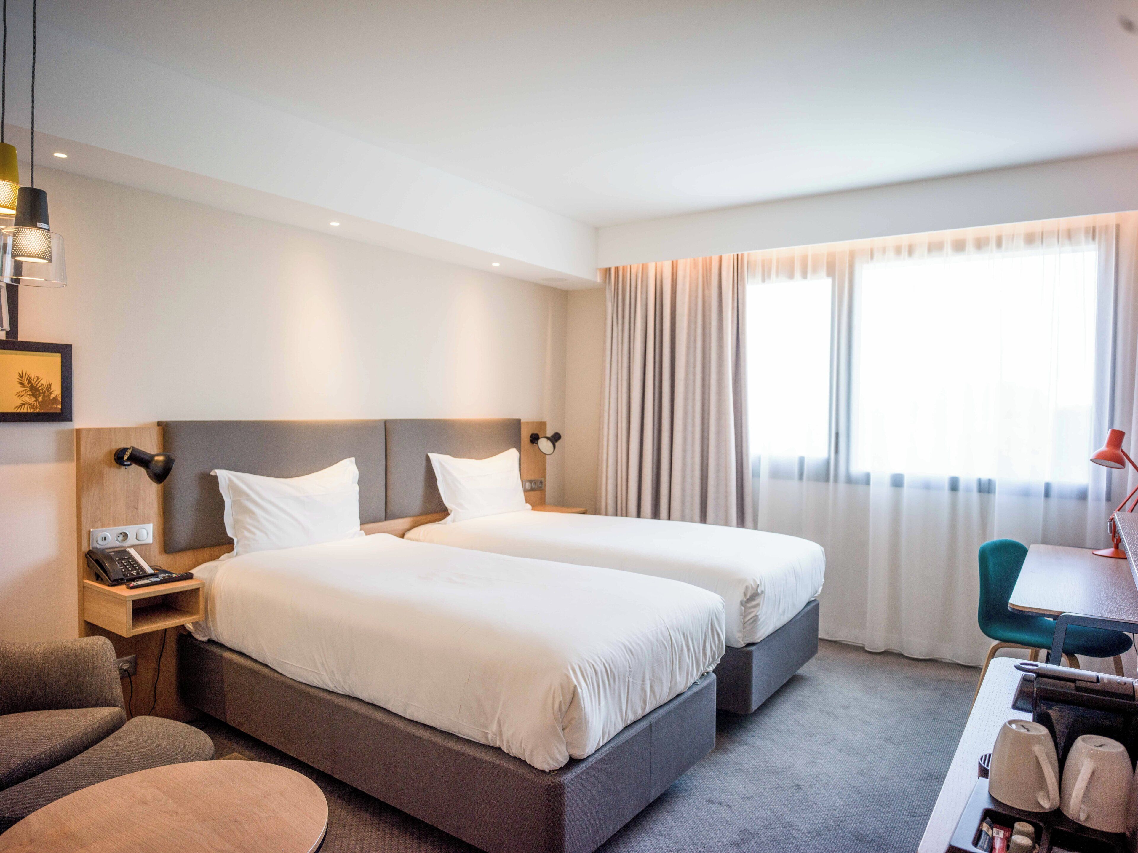 Ein Hotelzimmer mit zwei Betten, einem Schreibtisch mit Stuhl, einer kleinen Sitzecke, einer Schreibtischlampe, einem Telefon und einem Fenster mit Vorhängen.