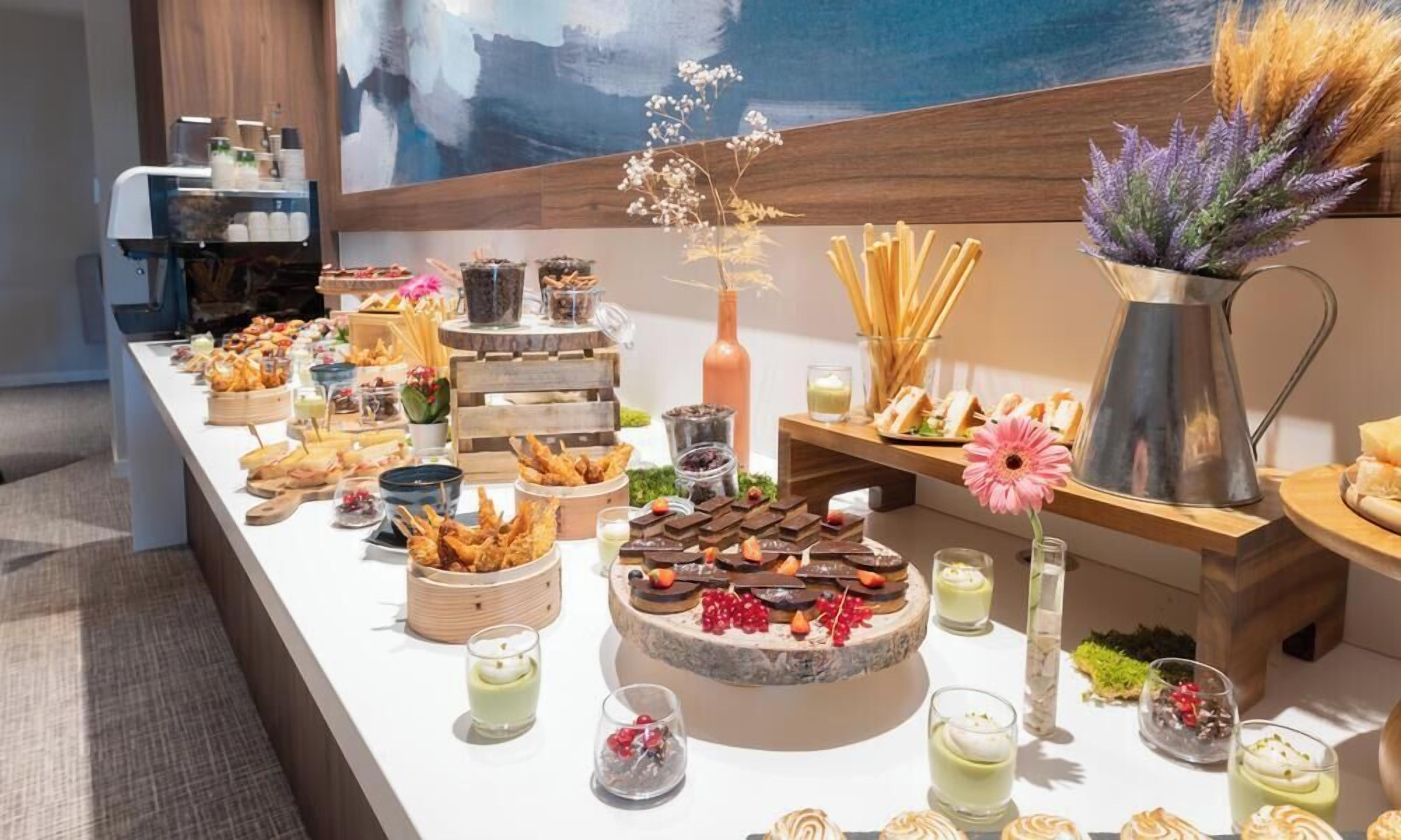 A buffet table with various food items and drinks, including pastries, desserts, and beverages, arranged on a white table with a modern design.