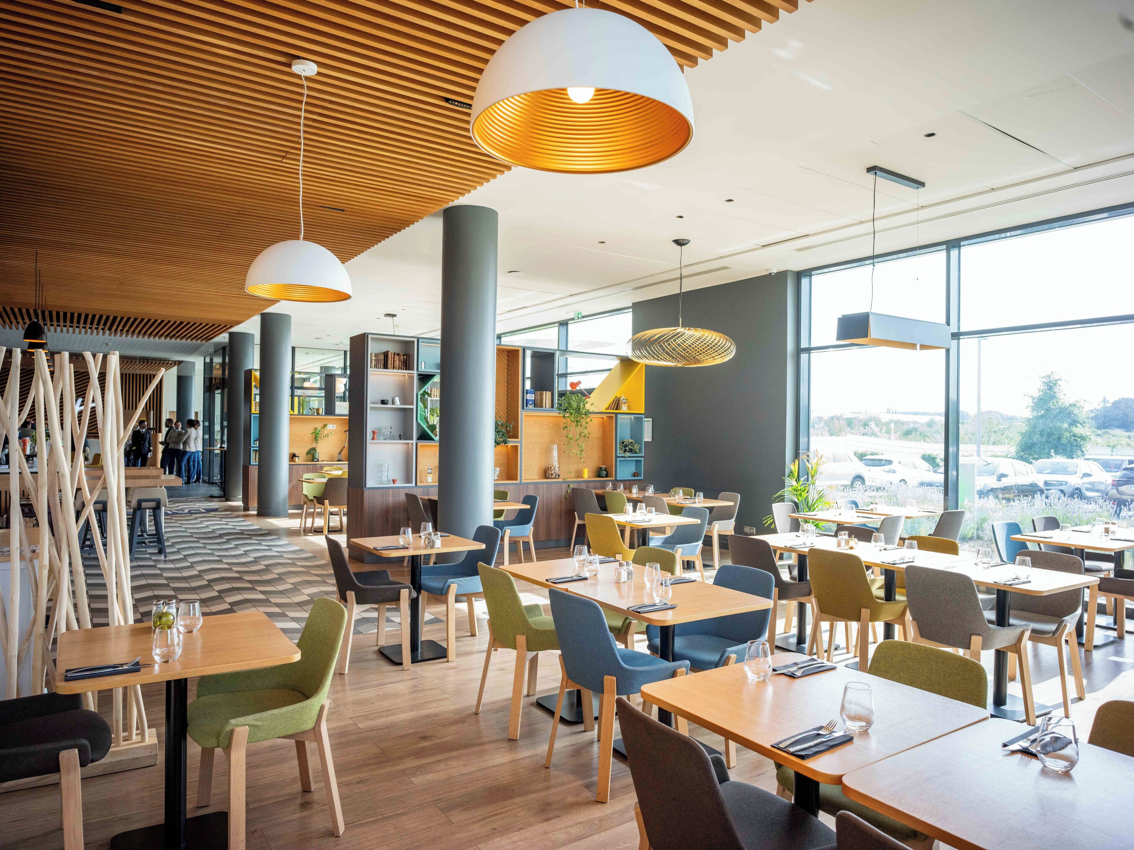 A modern restaurant interior with wooden ceiling panels, large windows, and a variety of seating arrangements.