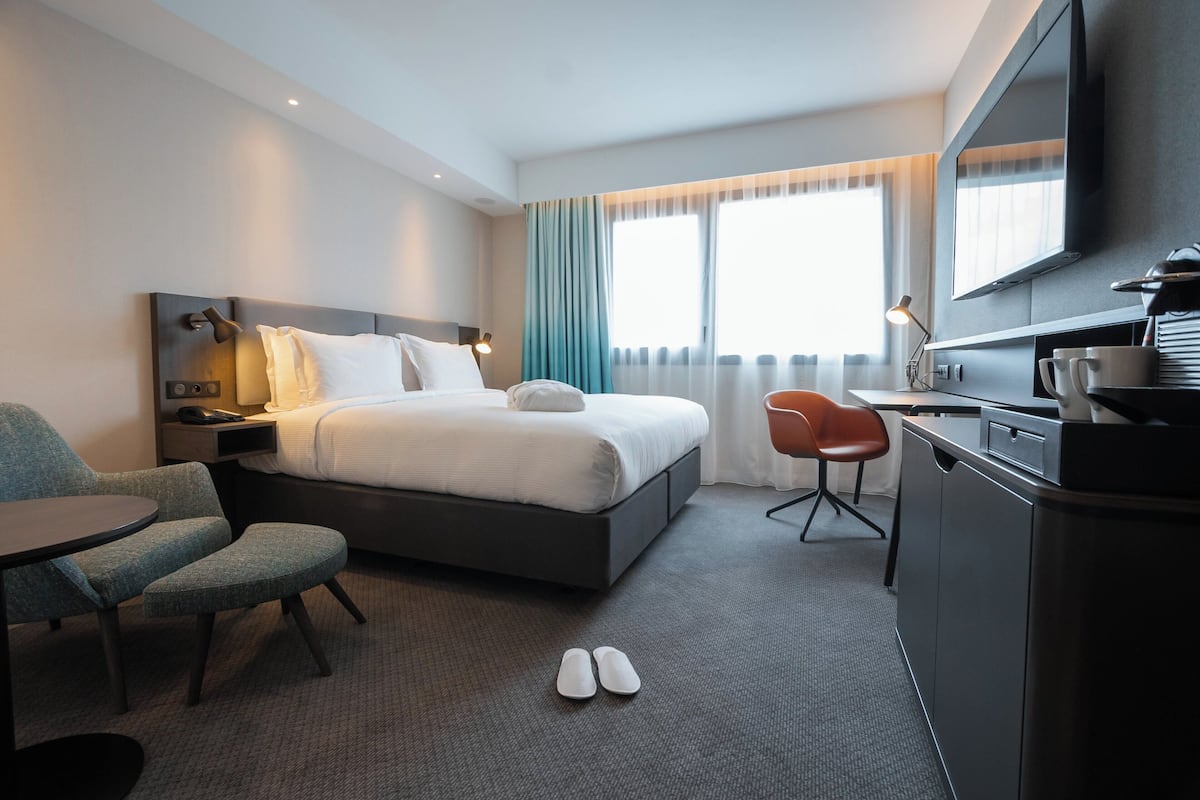A hotel room with a bed, a desk, a chair, a TV, and a window with curtains.