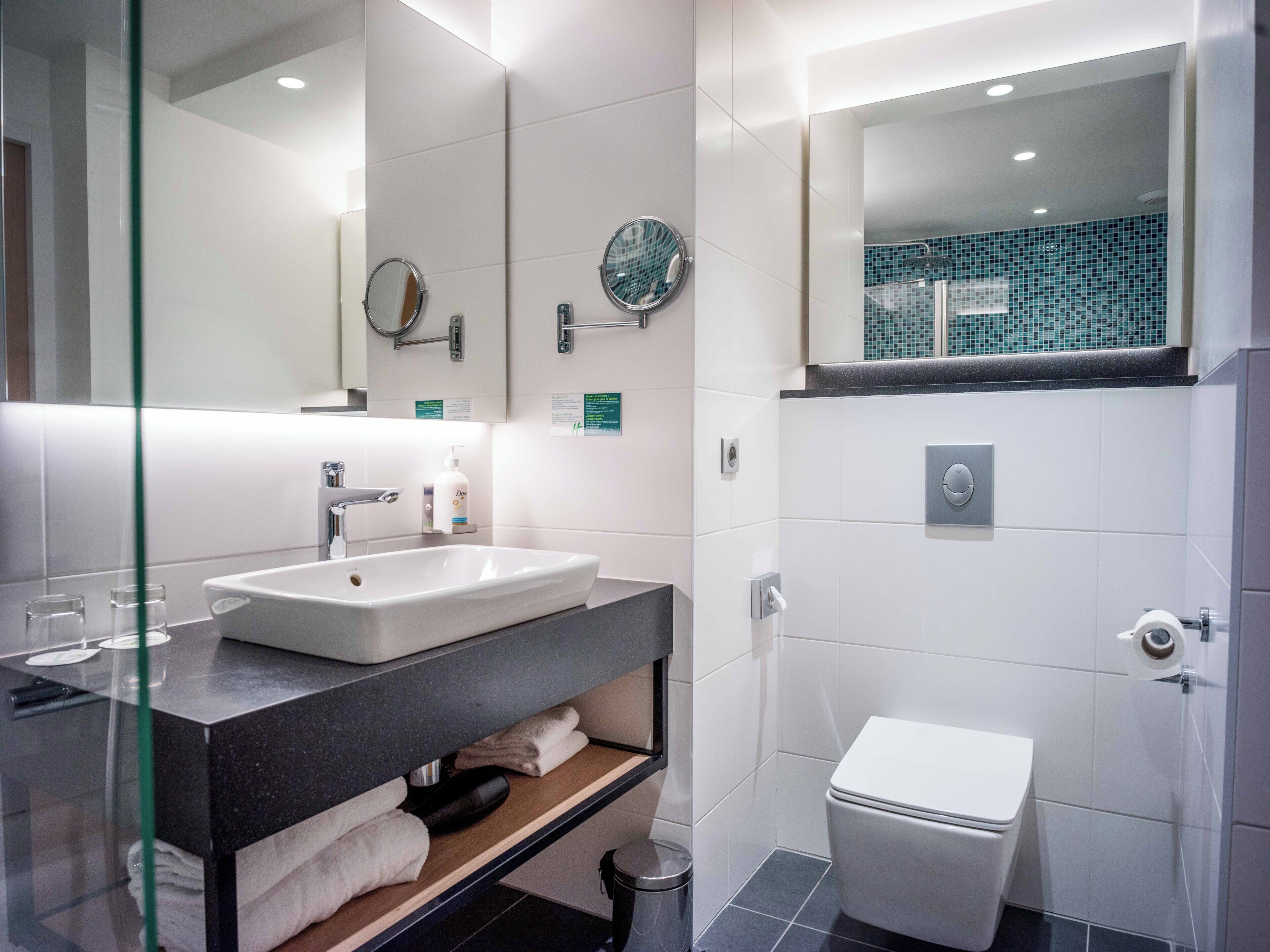 A modern bathroom with a white sink, a mirror, a toilet, and a shelf with towels.