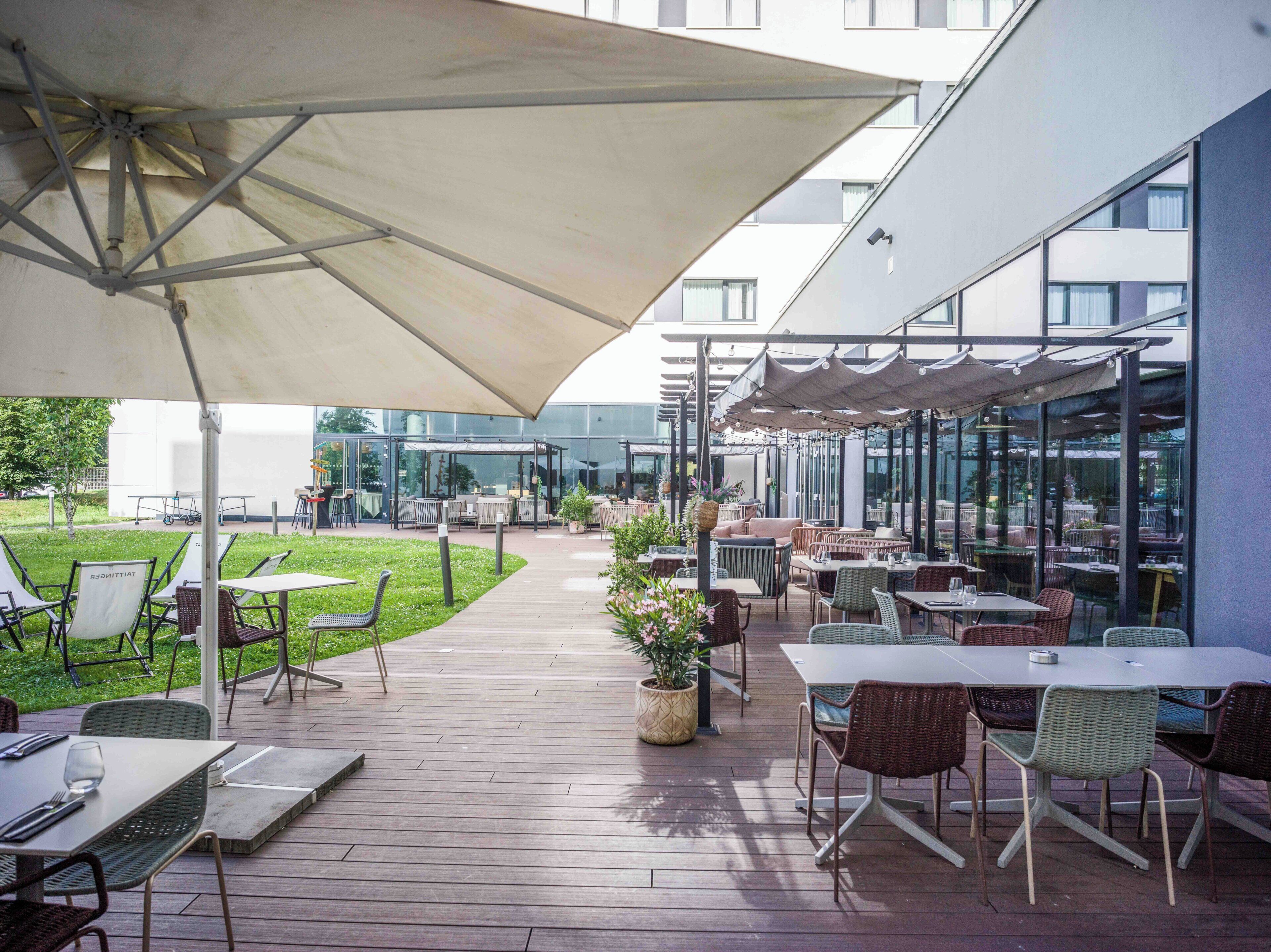 Eine Terrasse im Freien mit Tischen und Stühlen, ein überdachter Sitzbereich und im Hintergrund ein Gebäudeblick.