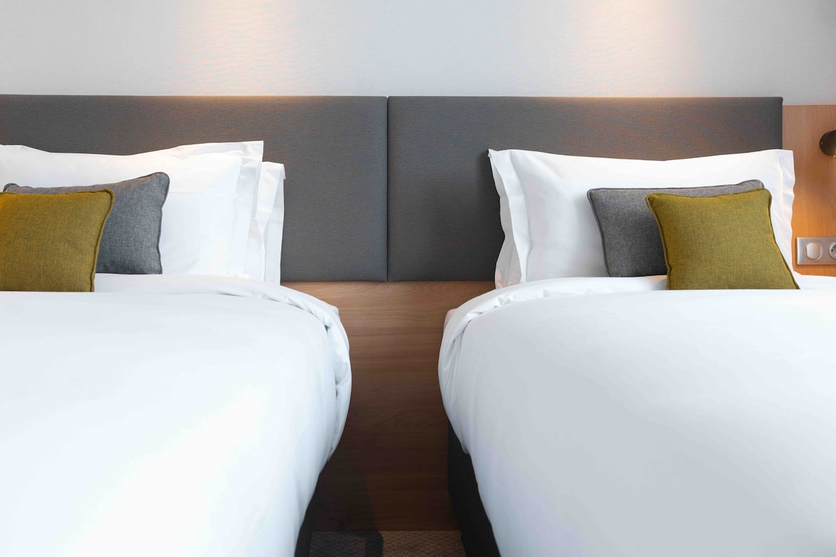 A hotel room with two beds, each with white bedding and a grey headboard. There are two wall-mounted lamps on either side of the beds.