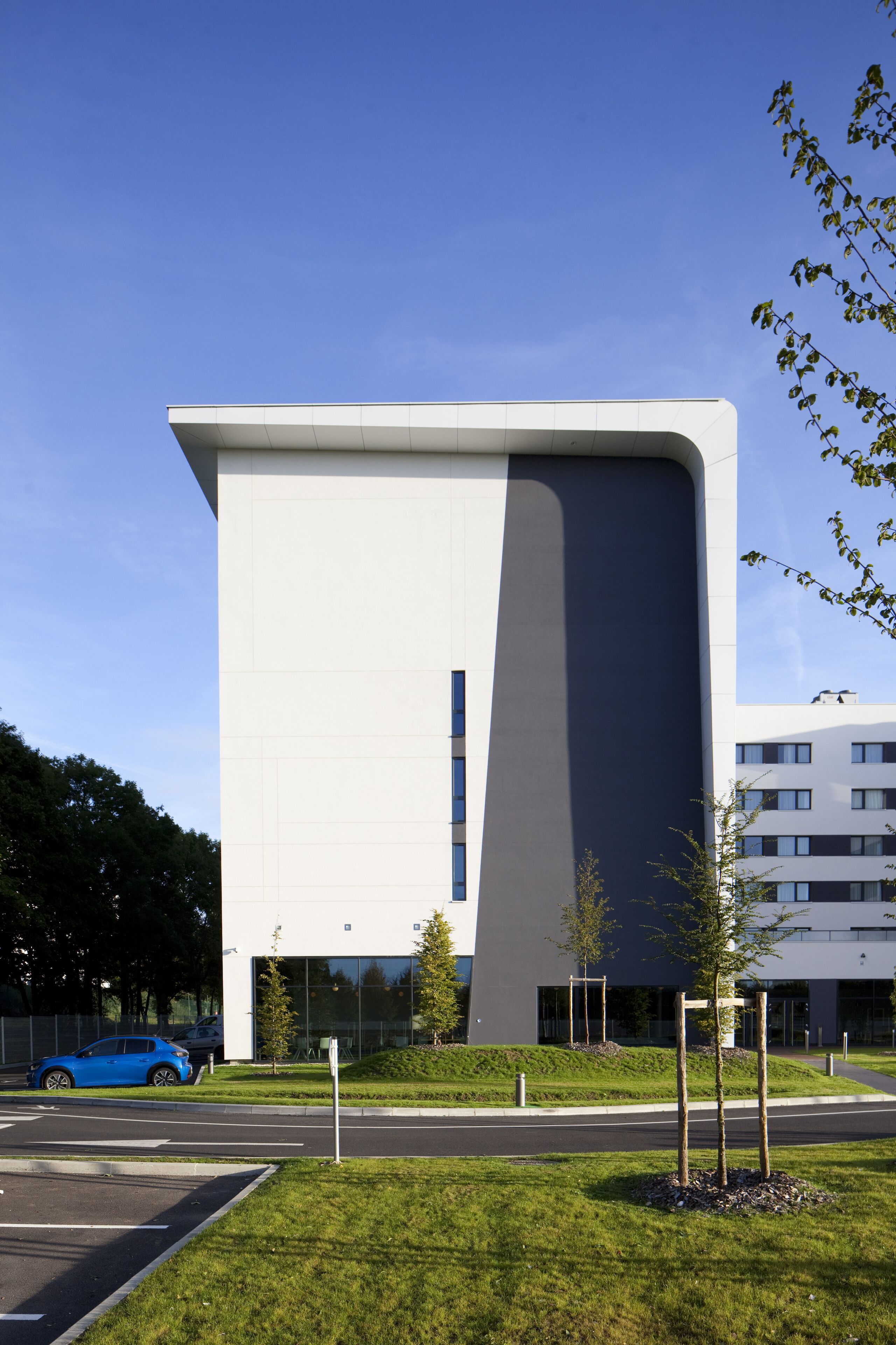 En moderne bygning med fladt tag, store vinduer og en parkeringsplads med en blå bil.