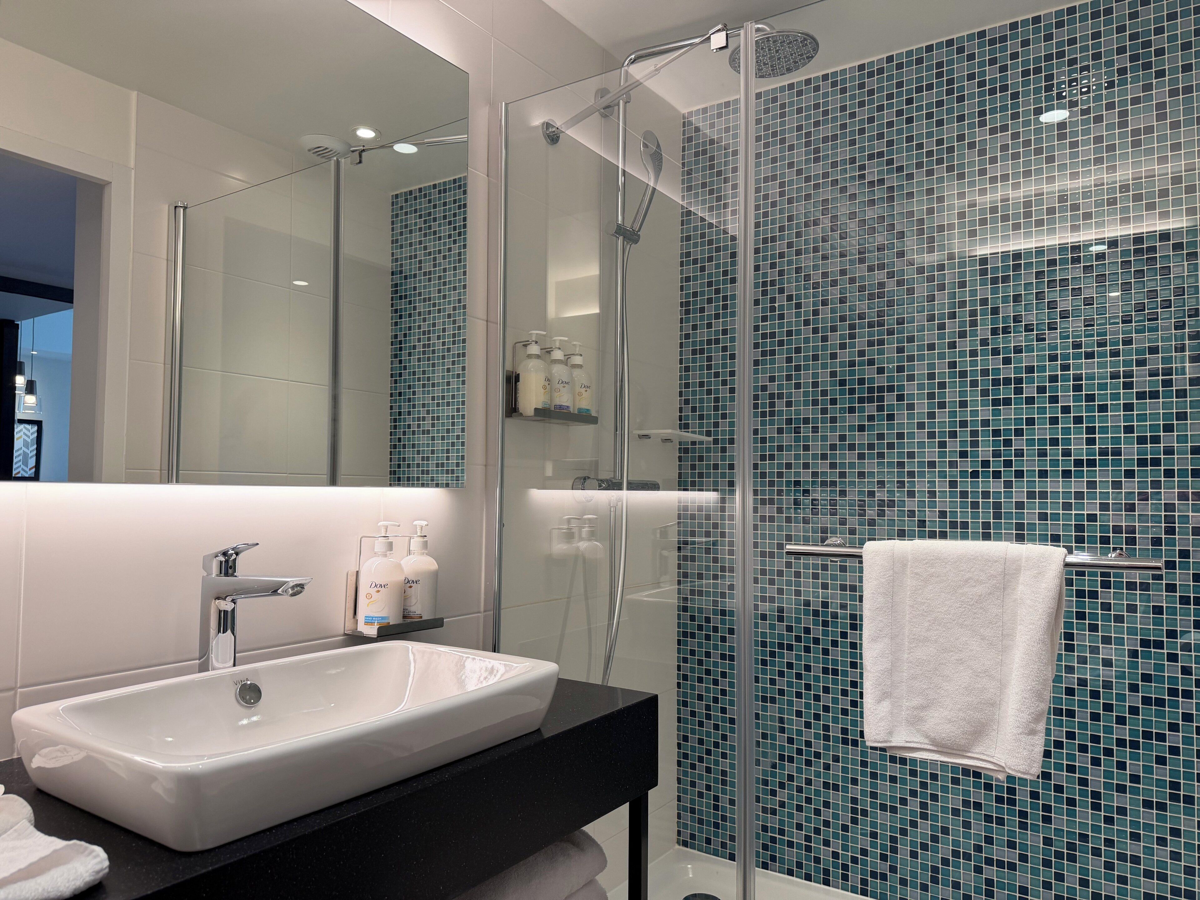 Un baño moderno con ducha de vidrio, lavamanos blanco y pared con diseño de mosaico azul.