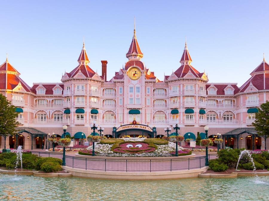 A large, pink, multi-story building with a central clock tower, surrounded by a fountain and greenery.