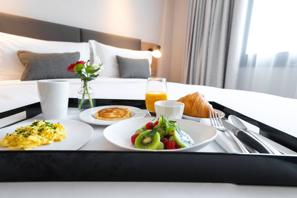 A breakfast tray with scrambled eggs, kiwi, raspberries, a croissant, orange juice, and a coffee cup on a bed with white linens and a gray headboard.