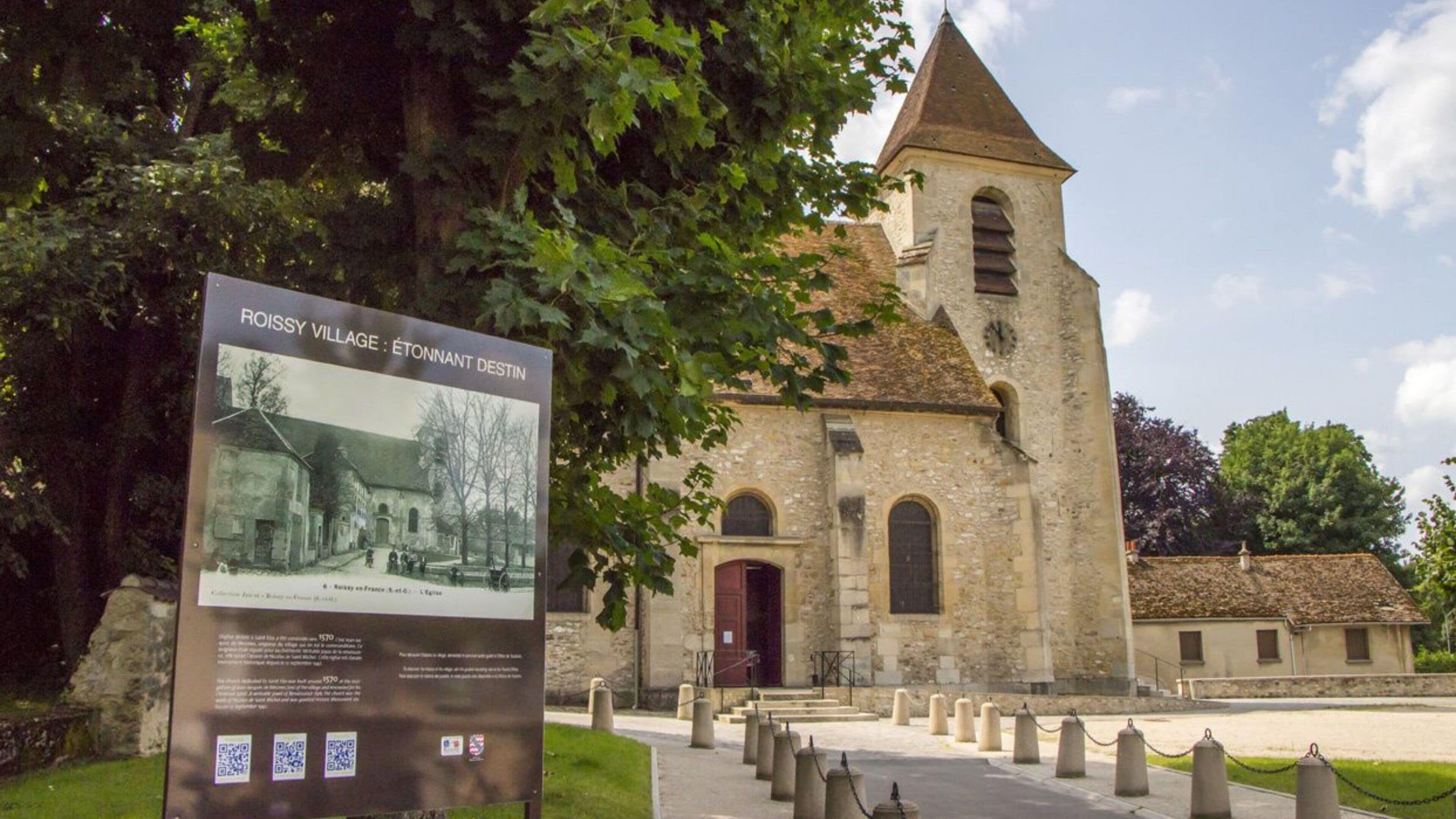 Una iglesia histórica con un campanario, rodeada de un jardín bien cuidado e indicación con información.