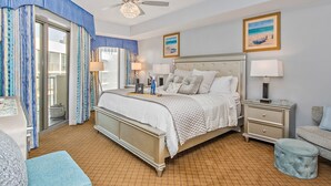 Une chambre à coucher avec un grand lit, deux tables de chevet, une commode, un ventilateur de plafond et une fenêtre avec des rideaux.