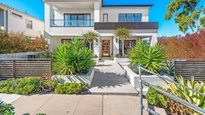 A modern two-story house with a central entrance, surrounded by a landscaped garden with various plants and a metal fence. - Luxury 4Bedroom home only 2 blocks from the beach! (Newport Beach)