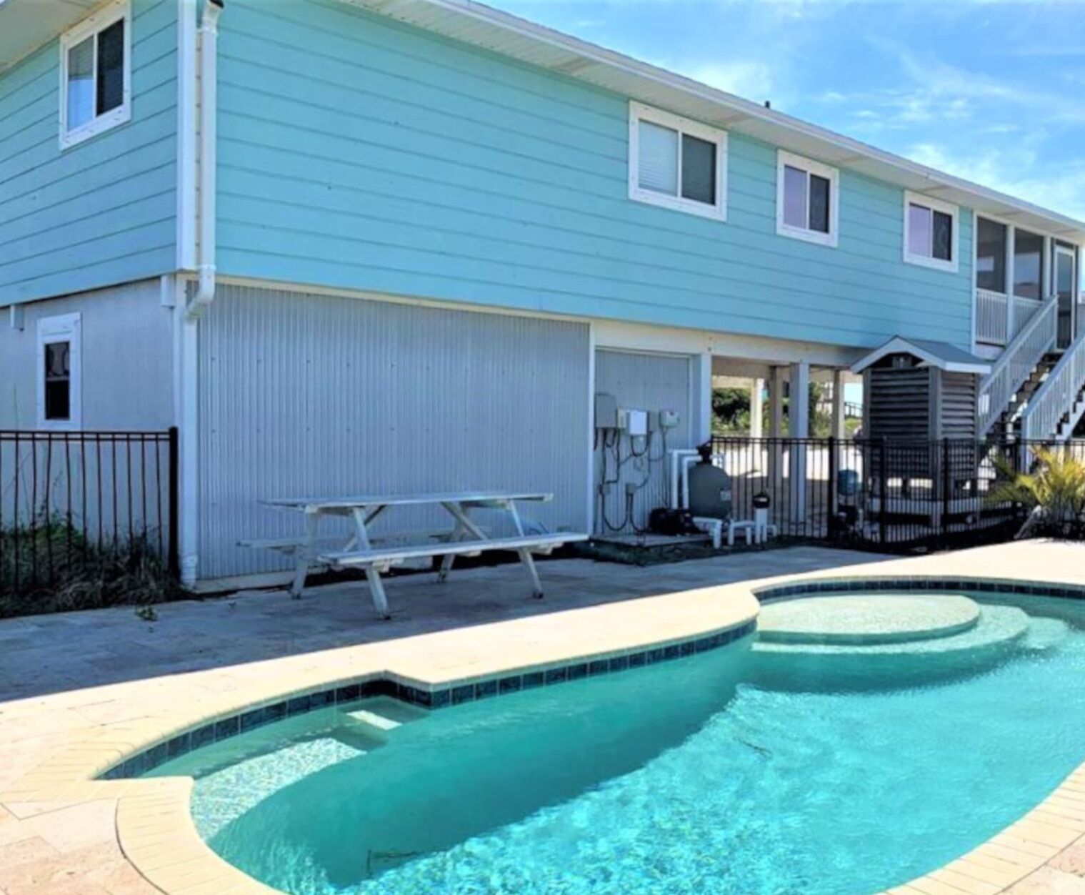 A pool area with a light blue building, a covered patio, and a staircase leading to an upper level.