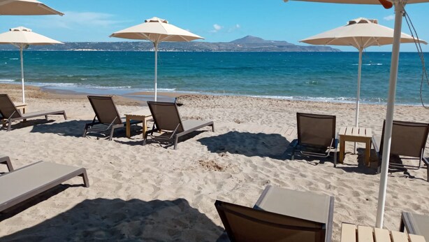 Una spiaggia con lettini e ombrelloni, vista libera sul mare e montagne in lontananza.