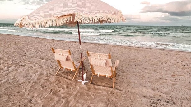 Een strand met twee rieten stoelen onder een gestreepte parasol.