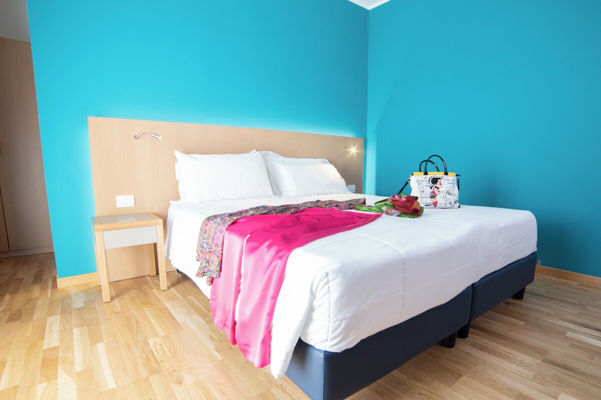 A neatly made bed with white linens and a pink blanket, a bedside table with a lamp, and a wall with a blue accent.